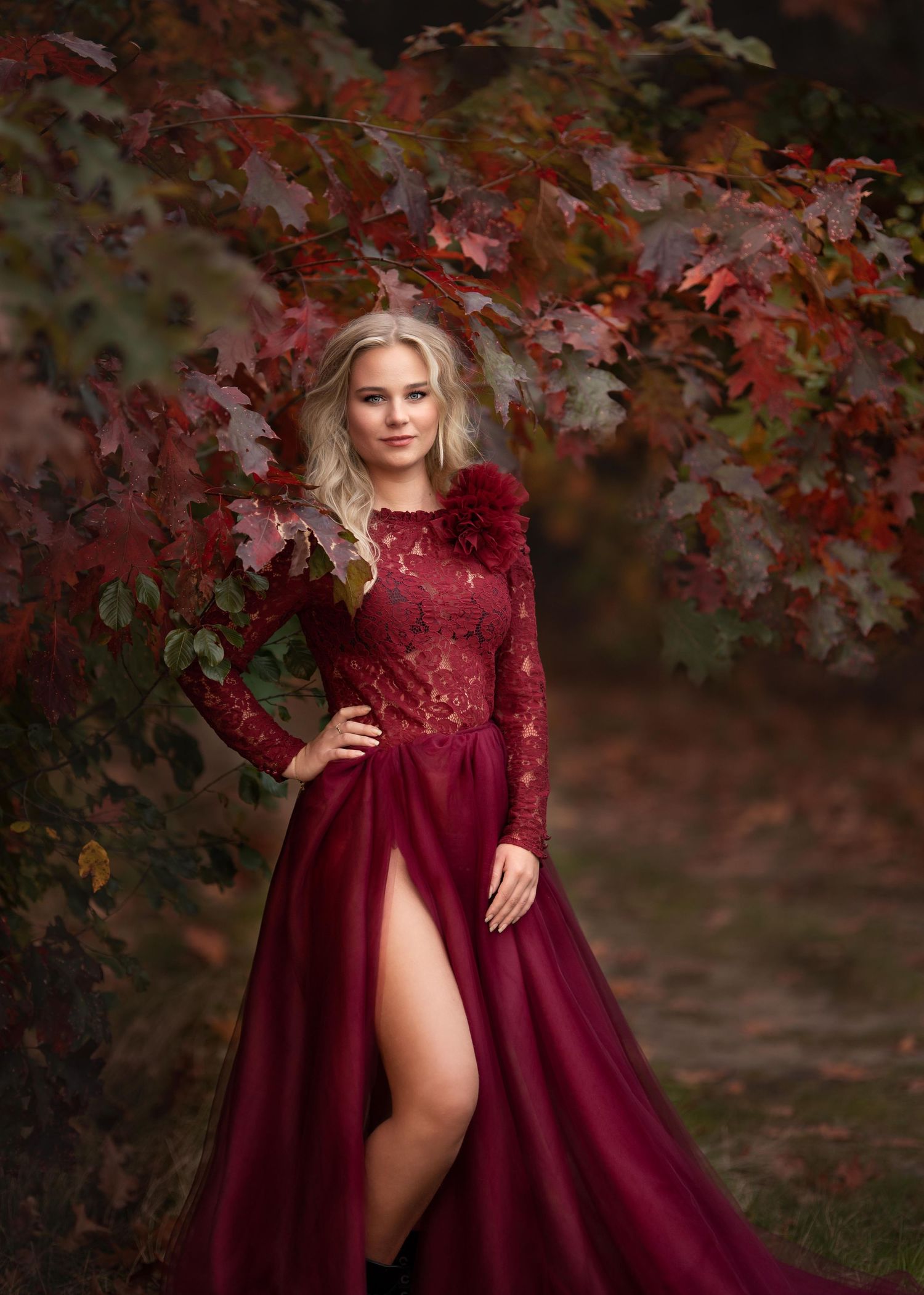 jonge blonde vrouw poseert zelfverzekerd in een rode glamourjurk voor een muur van herfstbladeren.