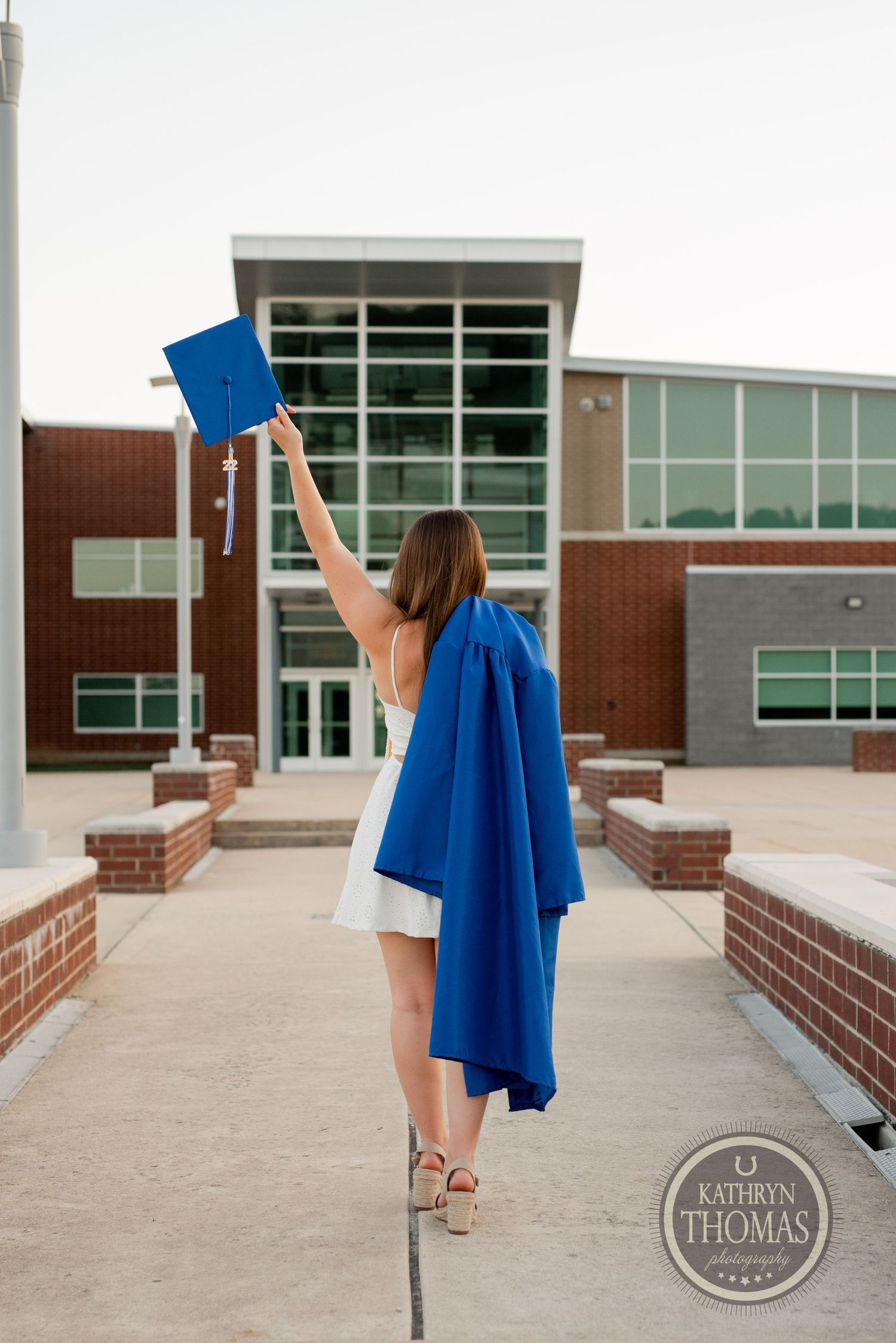 Graduate holding up blue diploma cover while wearing graduation regalia outside school entrance.