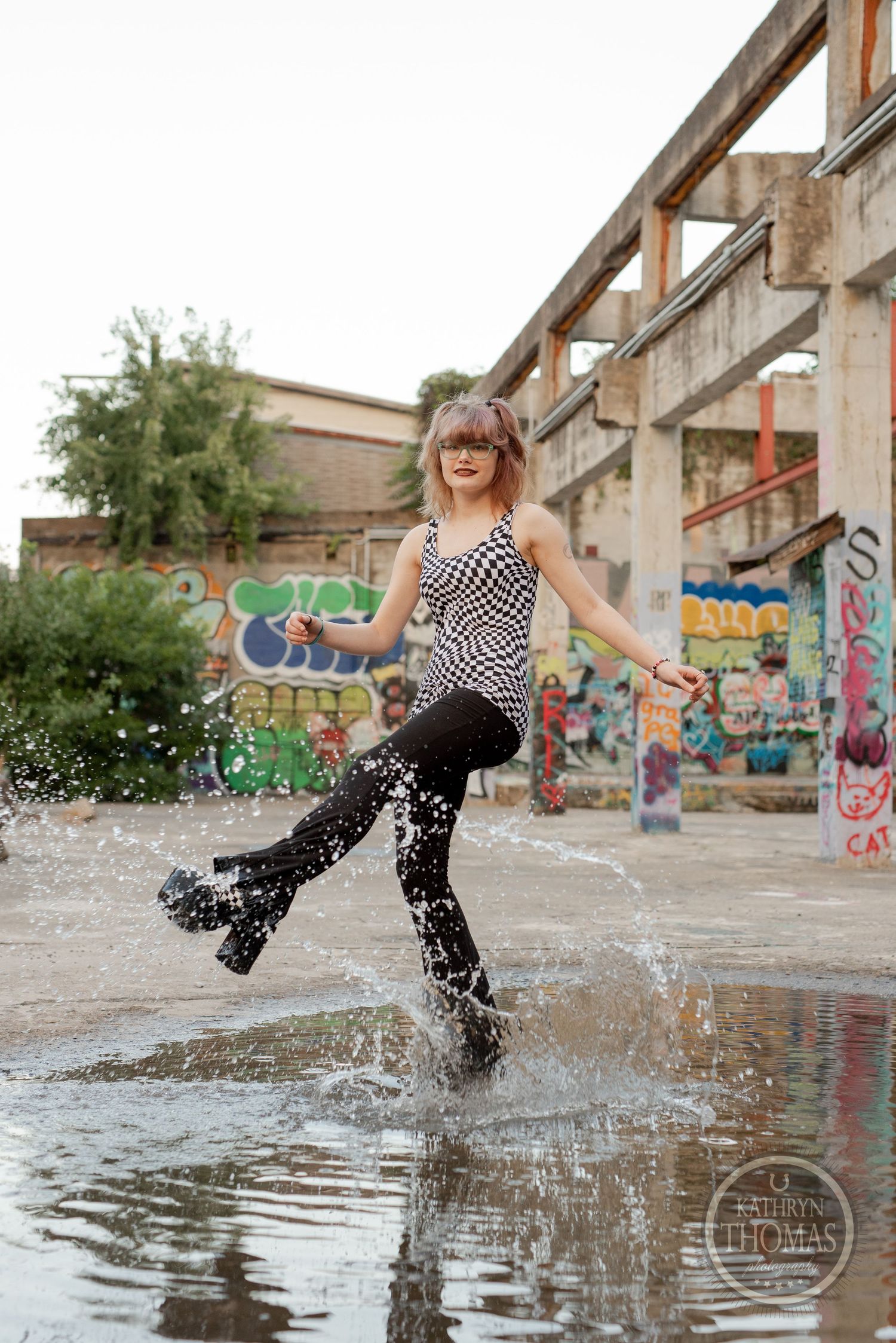 Playful splash photo in puddle wearing checkered top and patterned boots near graffiti wall.