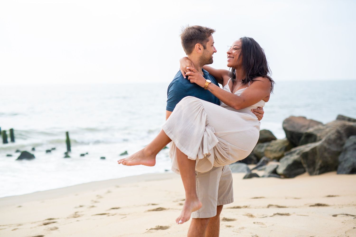 Romantic couple embraces on sandy beach with ocean waves and rocks in background.