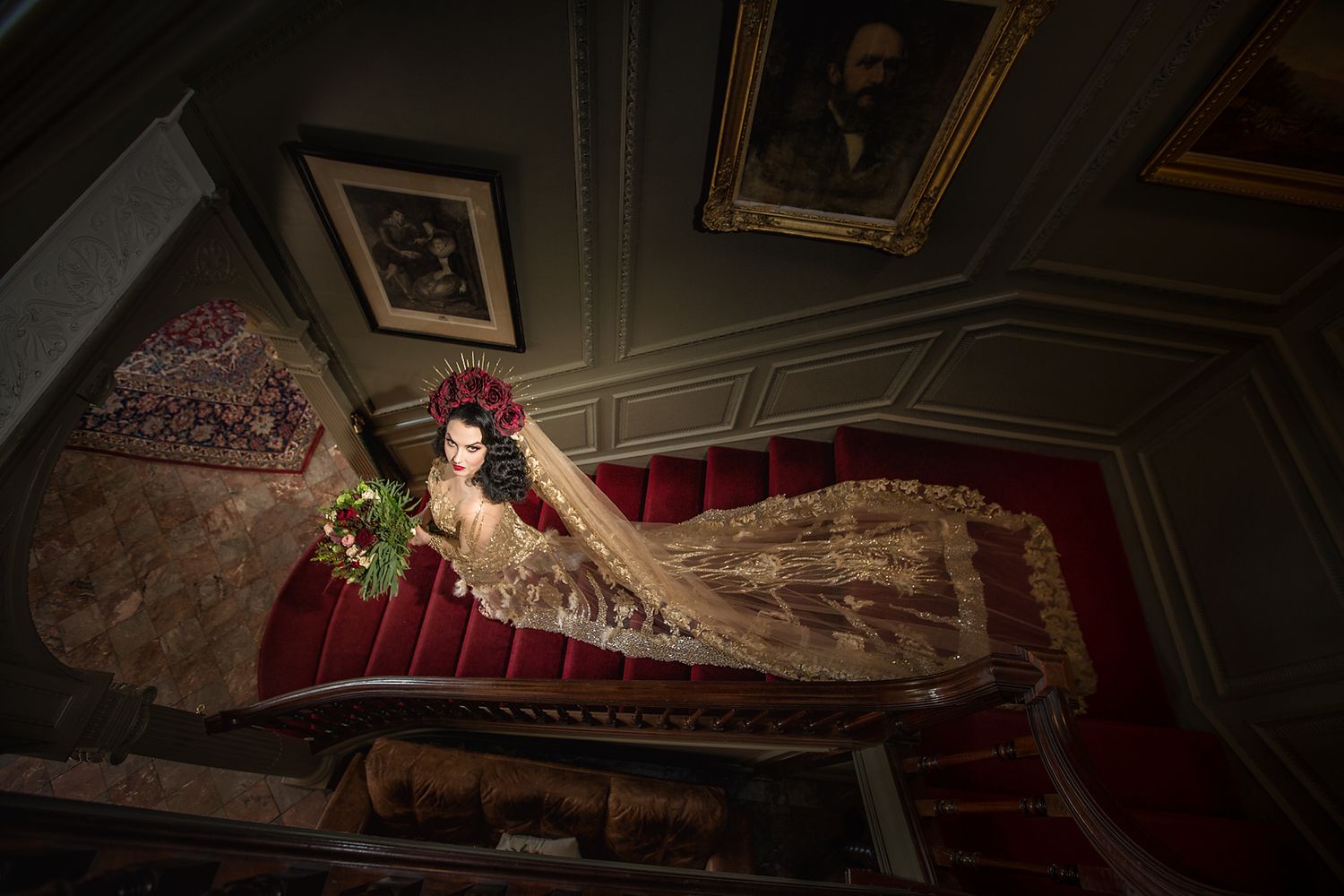 Gothic bride wearing a gold wedding dress walks down a red carpet staircase holding flowers at Thornton Manor