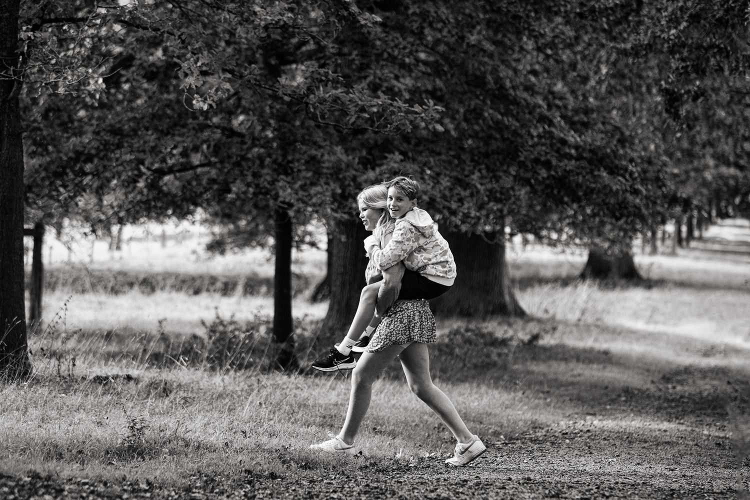 A young teenager carries a younger boy on her back and walk along a path surrounded by trees and tall grass