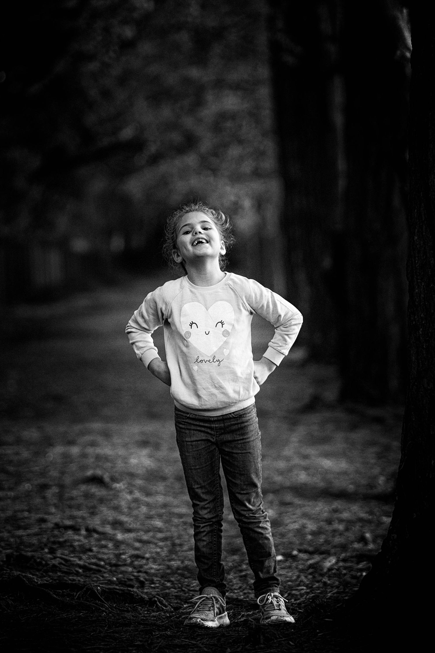 A young stands in a dark wood with her hands on her hips and smiles