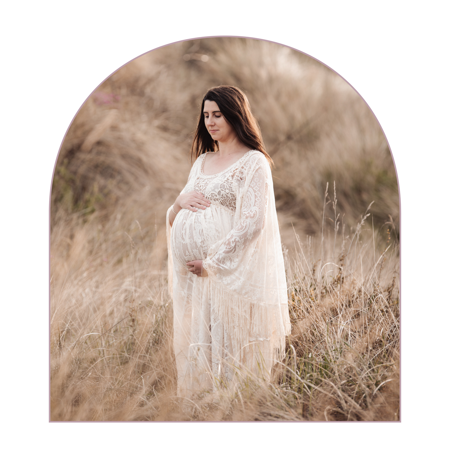 Soft maternity portrait in a delicate lace dress amidst tall grasses at sunset.