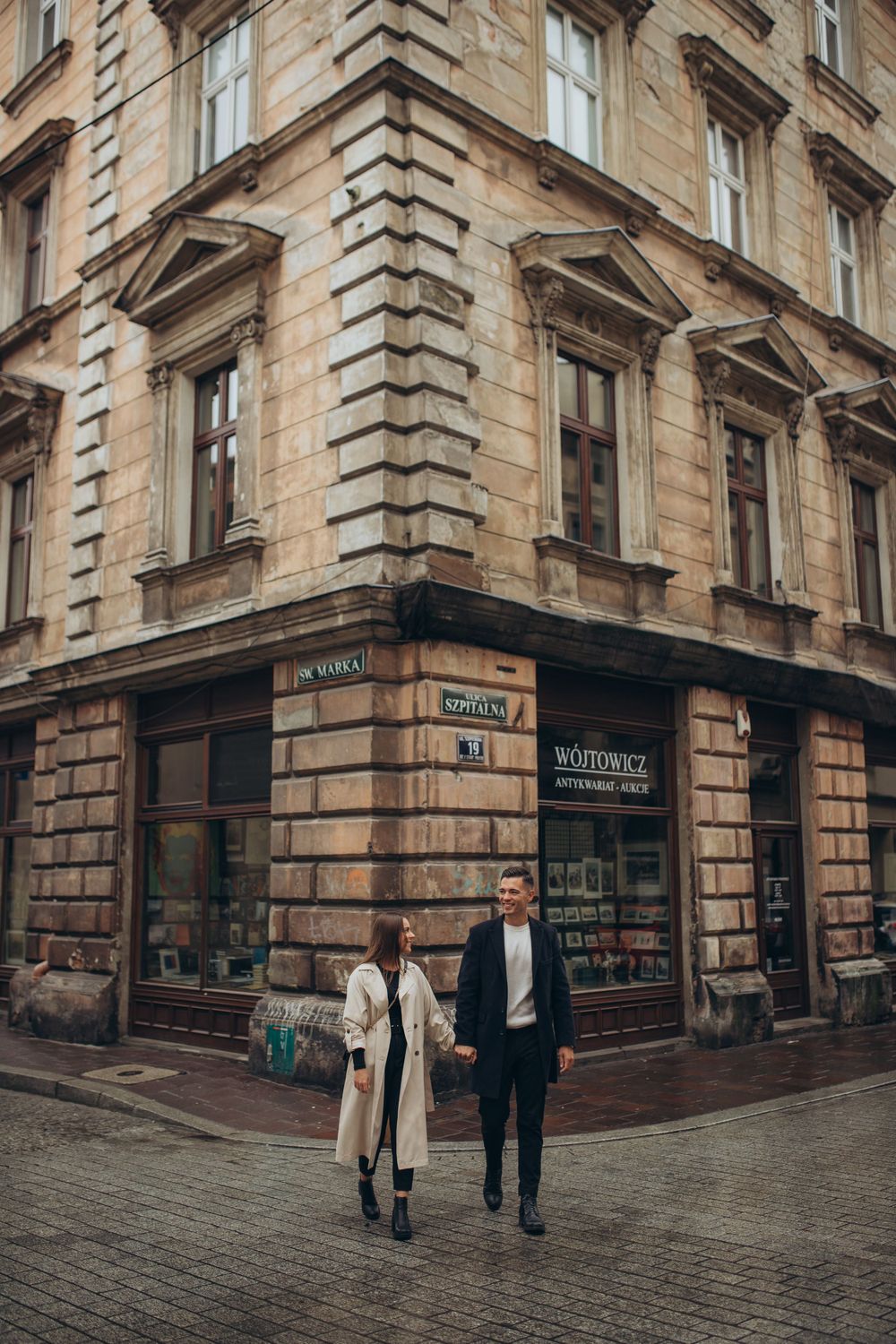 Two people walking together outside a vintage corner shop building with ornate stone architecture.