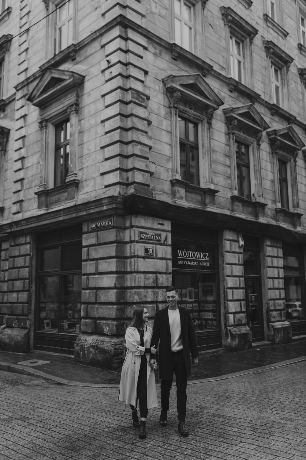 A couple stands in front of a historic stone building with a corner storefront on a cobblestone street.