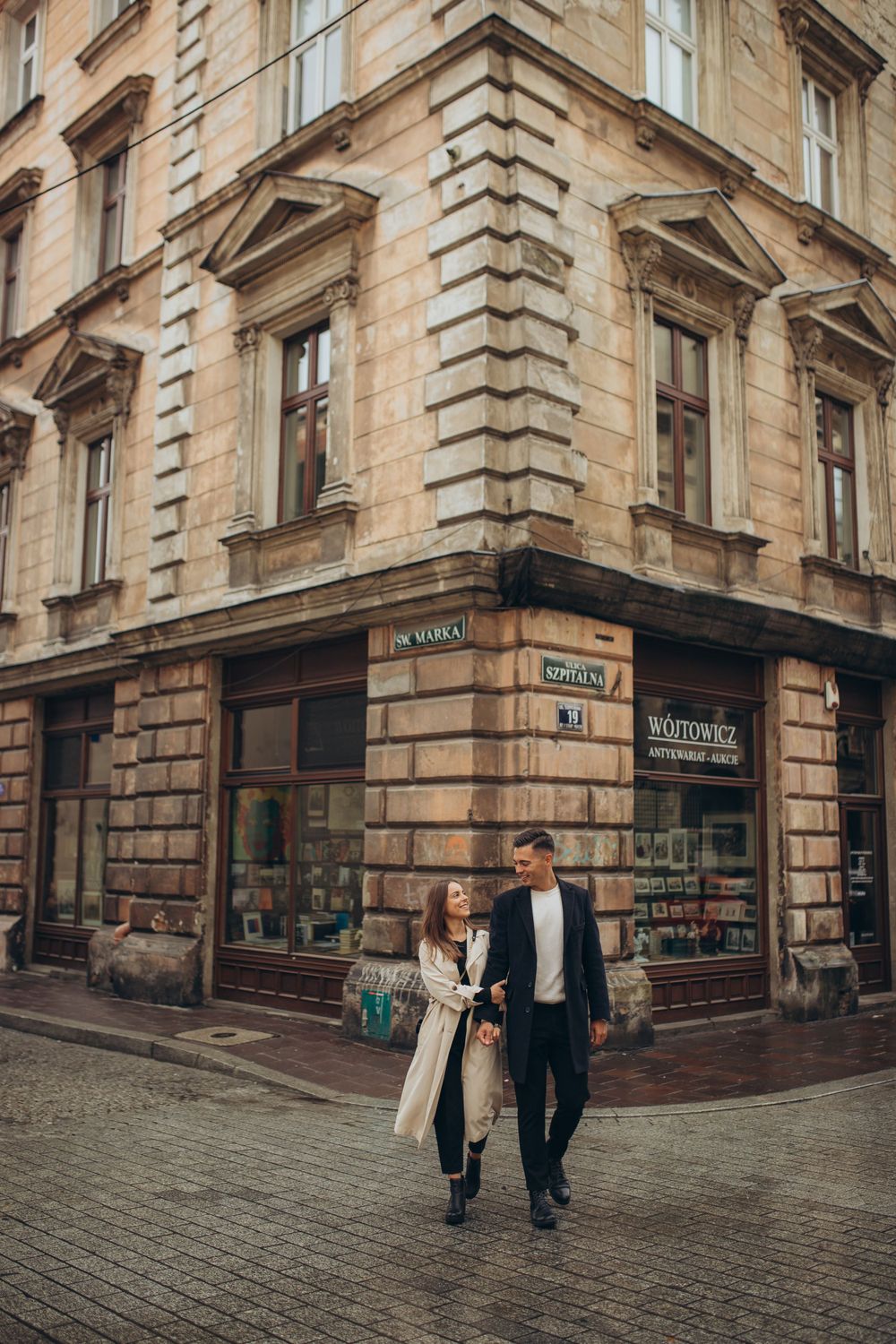A couple strolls past an old stone corner building with classic European architecture.