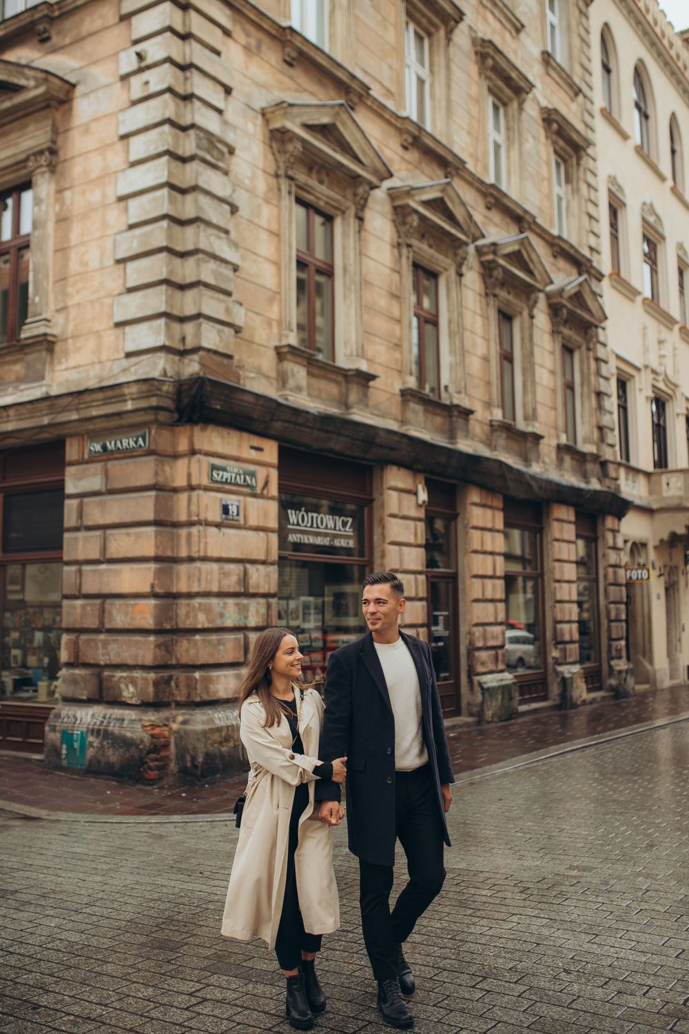 Two people walking on wet cobblestone street beside historic stone building in city center.