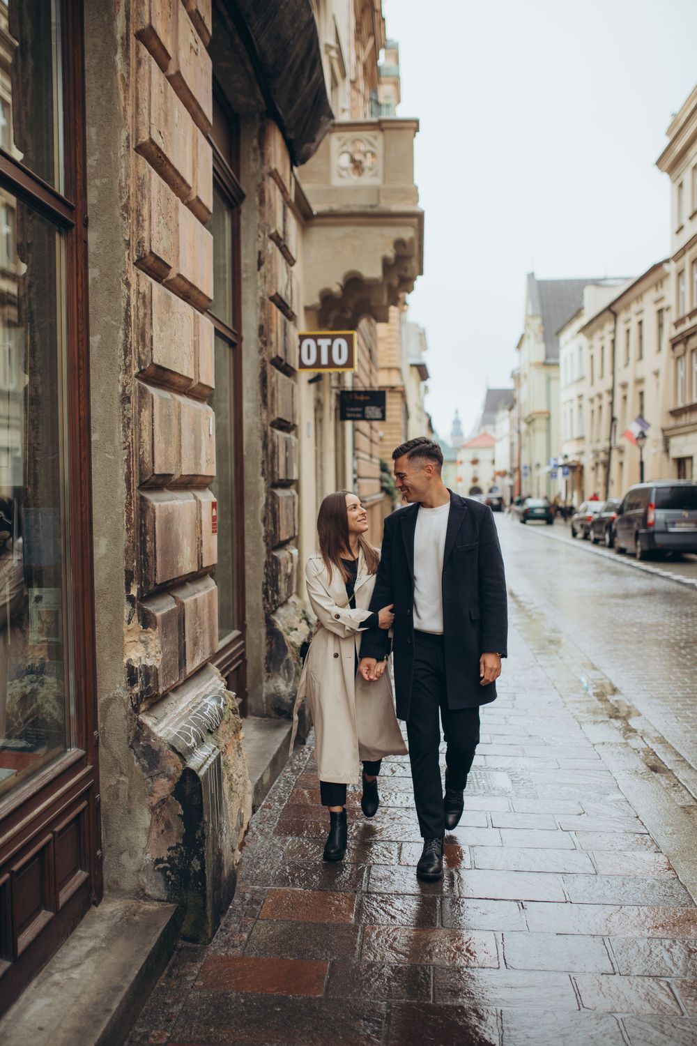 Two people walking together on wet sidewalk past historic stone buildings and parked cars.