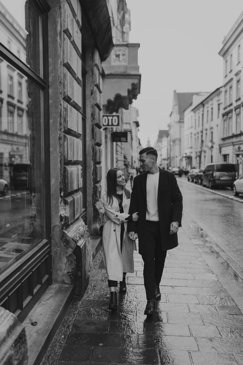 A couple walking down a rainy street past historic storefronts in black and white.