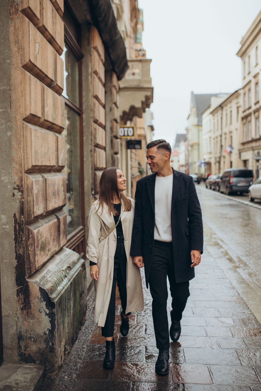 Two people walking and talking on wet cobblestone street beside historic architecture.