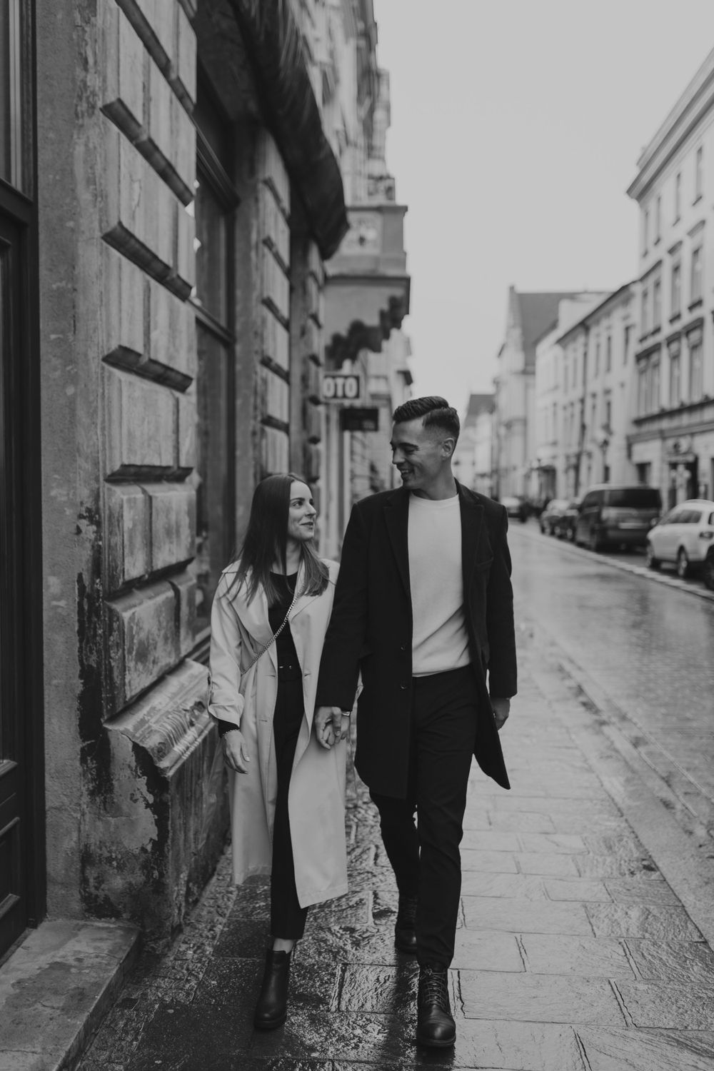 A couple shares a romantic moment while walking down a rainy European street in black and white.