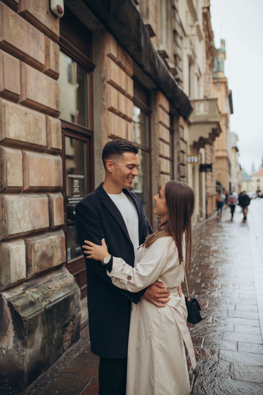 Two people embrace while sharing a happy moment outside historic stone buildings on wet street.