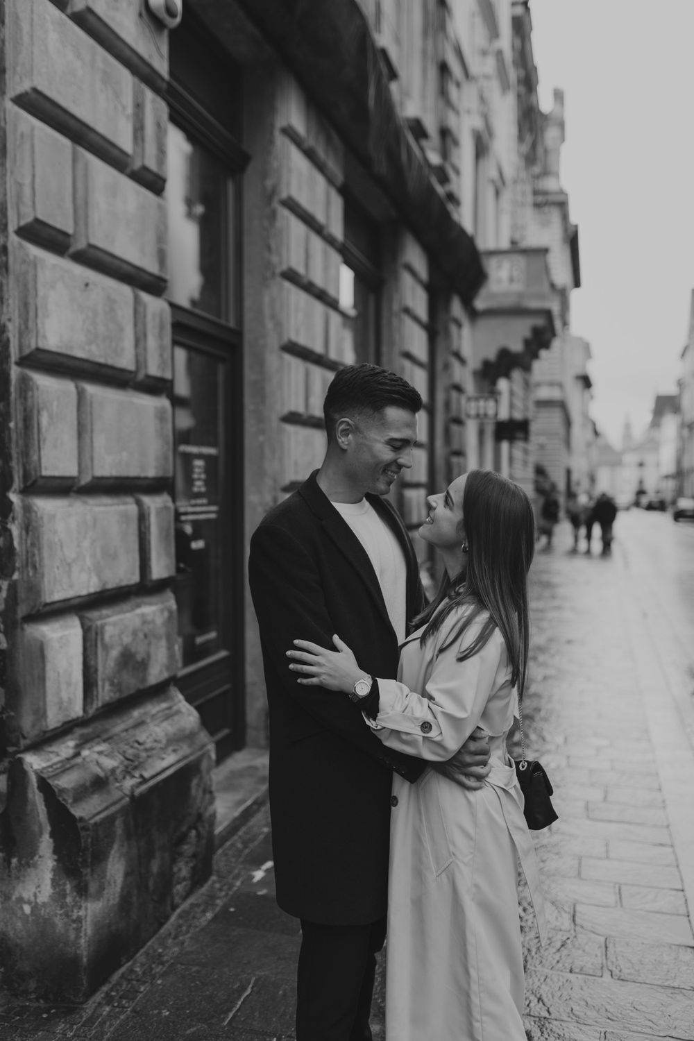 A romantic couple embraces on a historic European street with classic architecture in black and white.