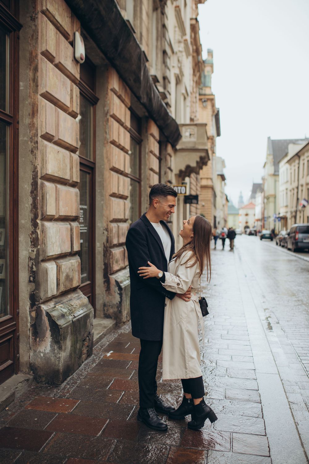 A couple shares an intimate moment against a stone building on a rainy cobblestone street.