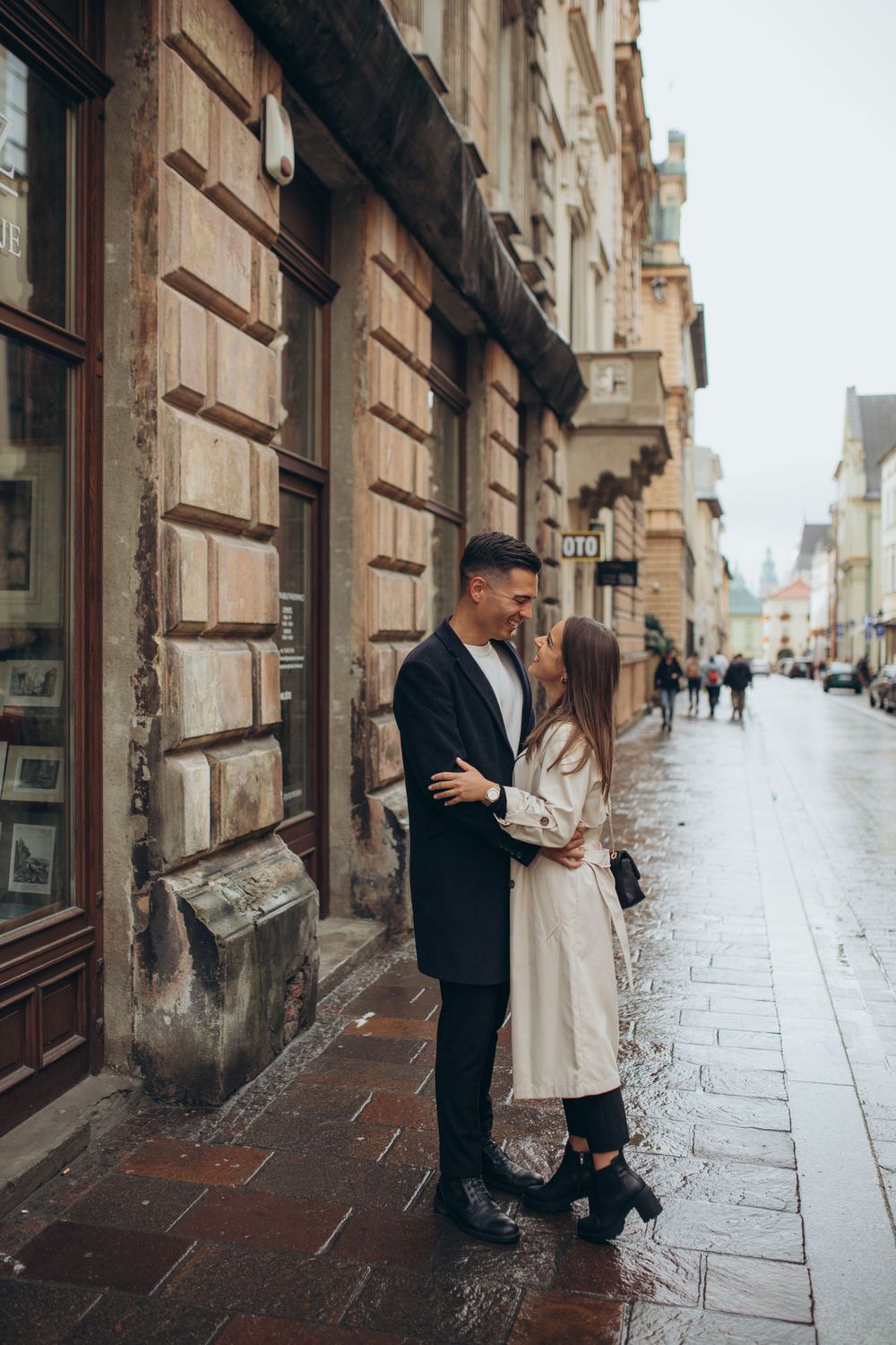 A couple in stylish coats embrace on a wet cobblestone street in front of historic architecture.
