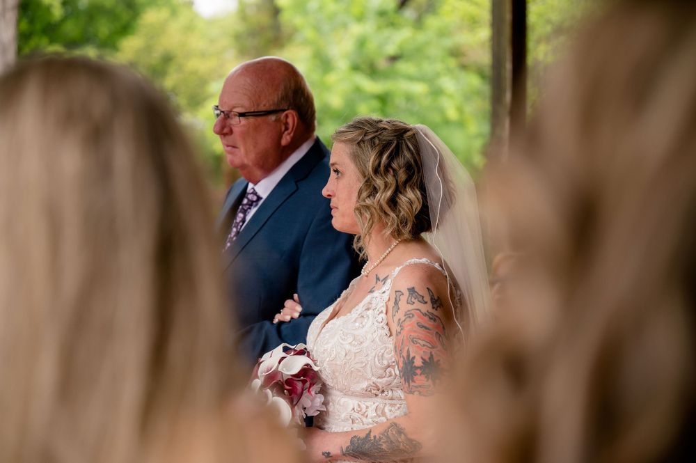 Bride in lace dress stands with companion during outdoor wedding ceremony against green foliage background.