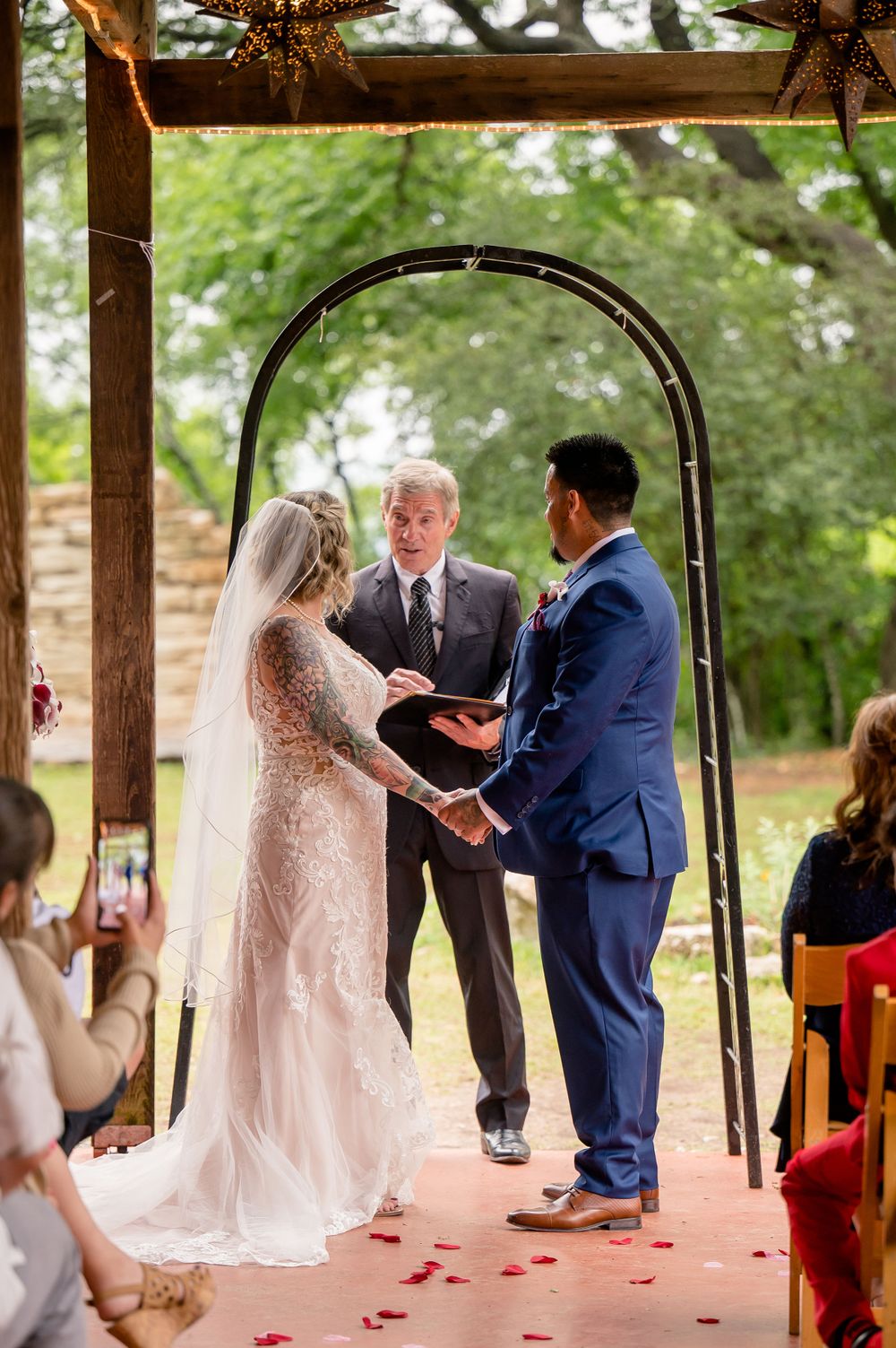 Outdoor wedding ceremony under wooden arch with officiant speaking to couple.