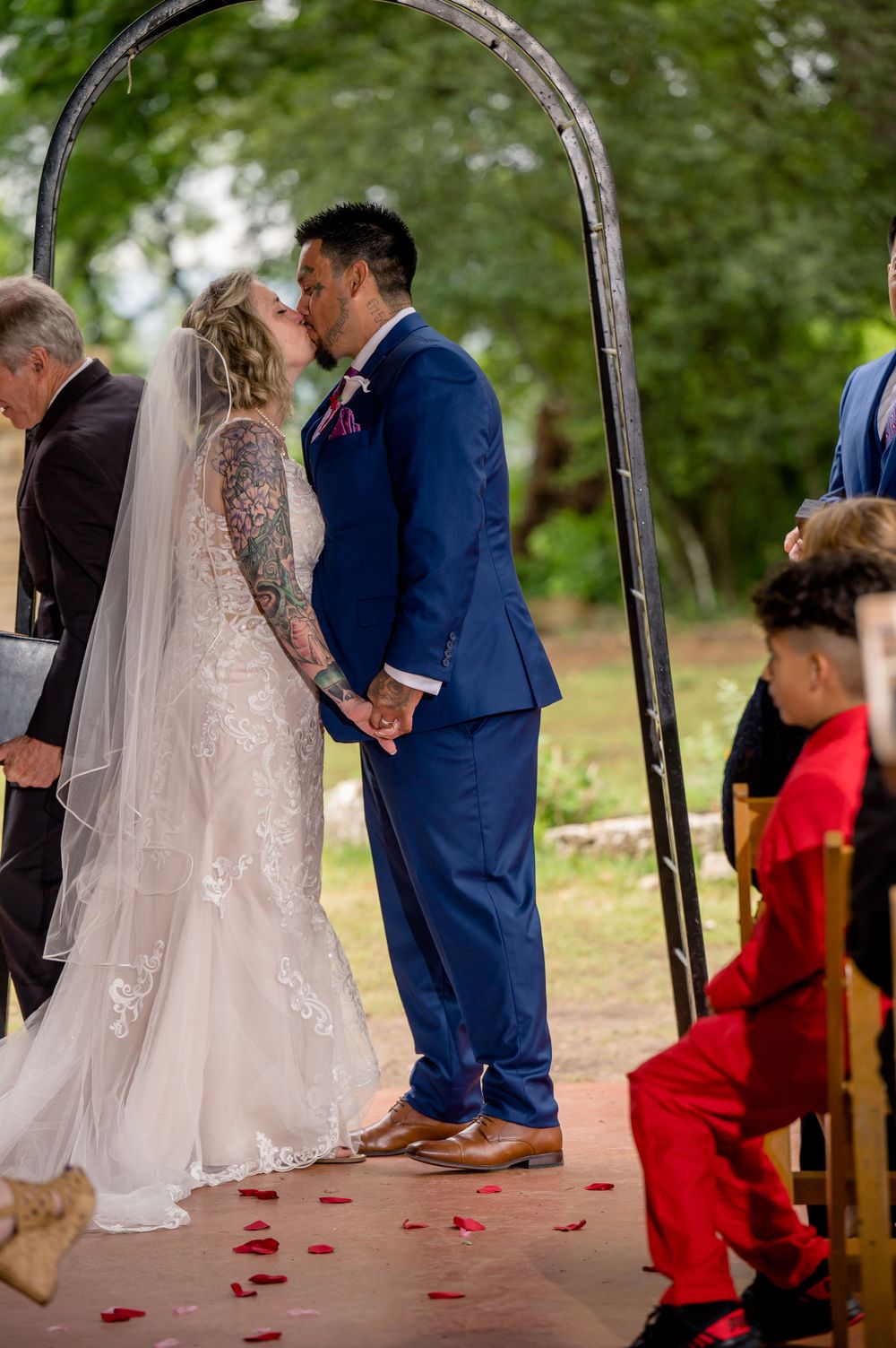 Wedding ceremony under rustic arch with scattered red petals on outdoor pathway.