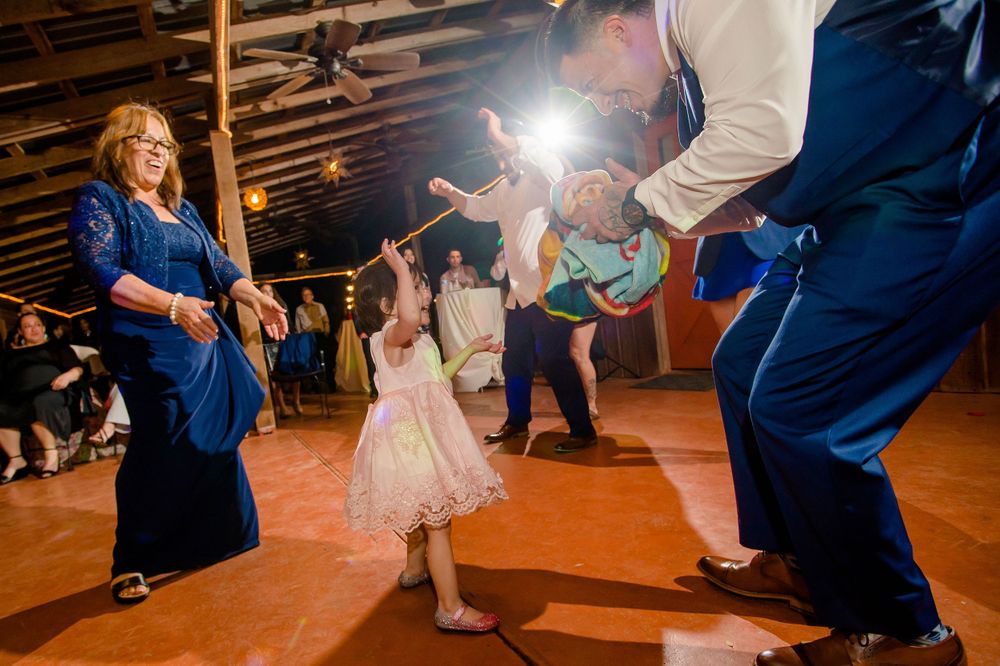 Young girl in pink dress dances with wedding guests in blue attire on rustic dance floor.