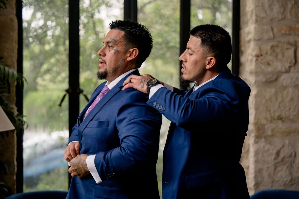 Two groomsmen in navy suits adjust each other's attire before wedding ceremony.