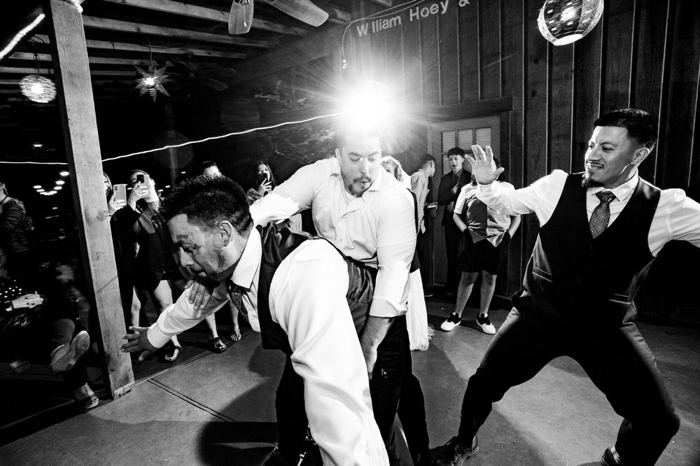 Black and white photo of guests dancing energetically at wedding reception in rustic venue.