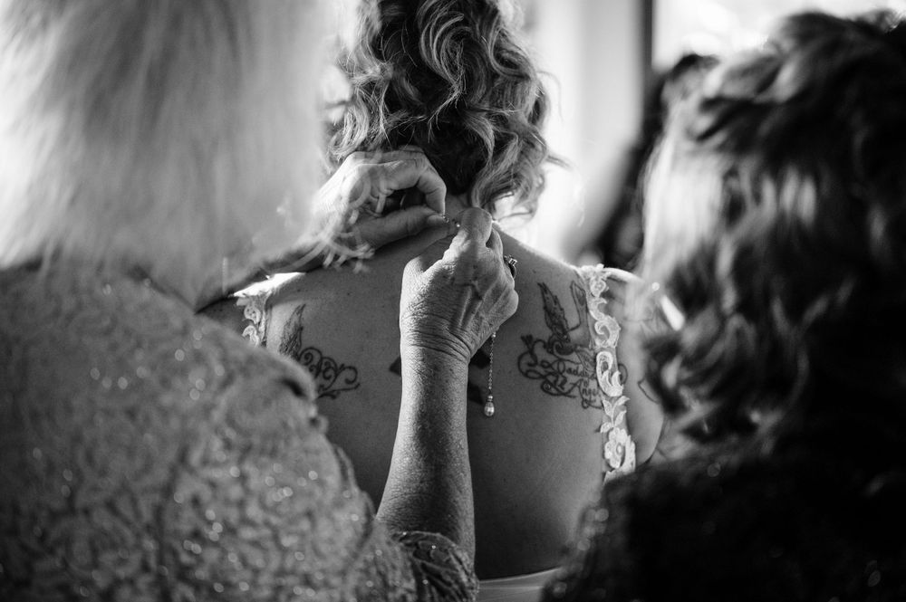 Black and white image of wedding dress preparation showing detailed back tattoo through sheer fabric.
