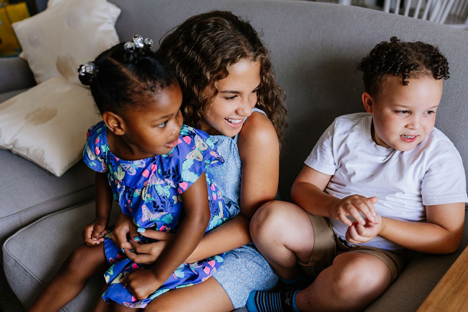 Three children sitting close together on a grey couch at home.