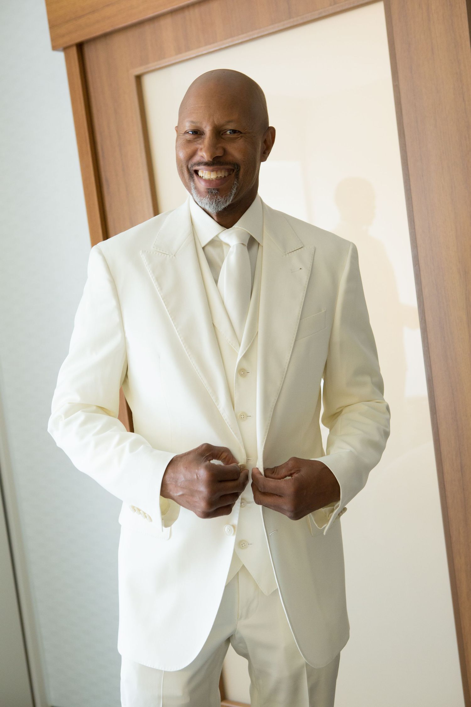 Groom in crisp white three-piece suit adjusts jacket while standing in doorway.