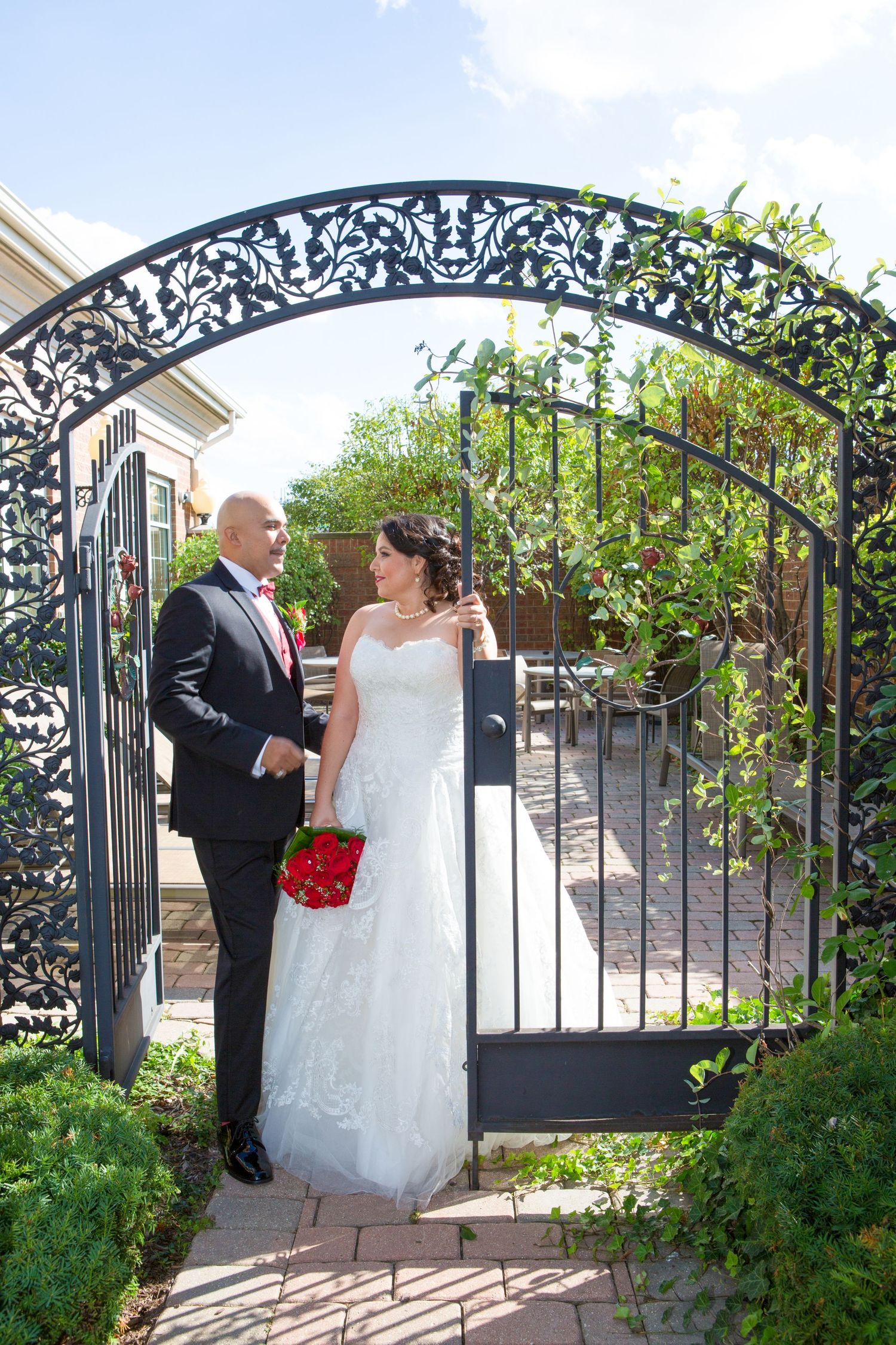A couple poses under an ornate iron archway with red roses in a garden setting.
