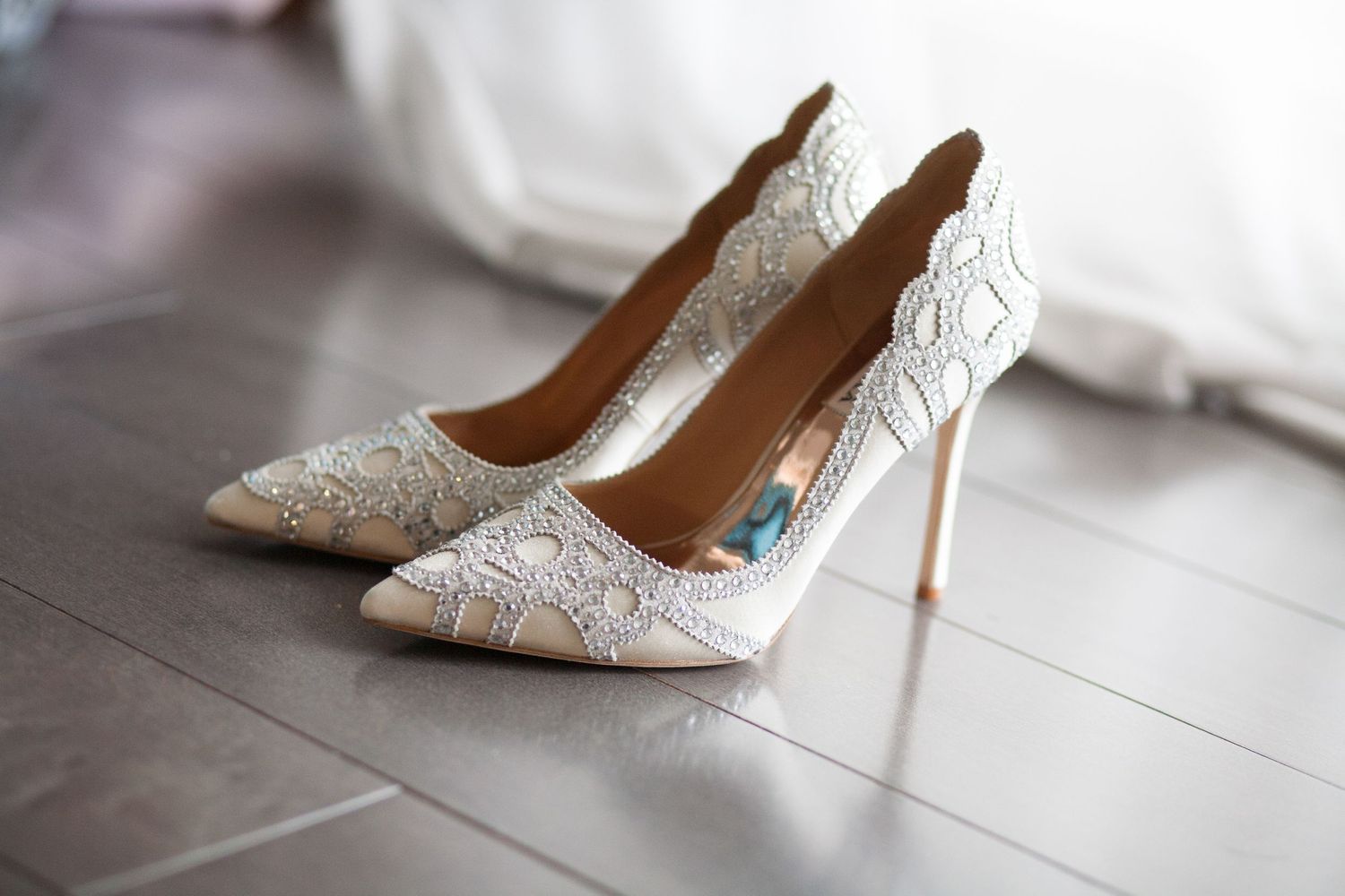 Elegant white lace bridal shoes with high heels displayed on a gray floor.