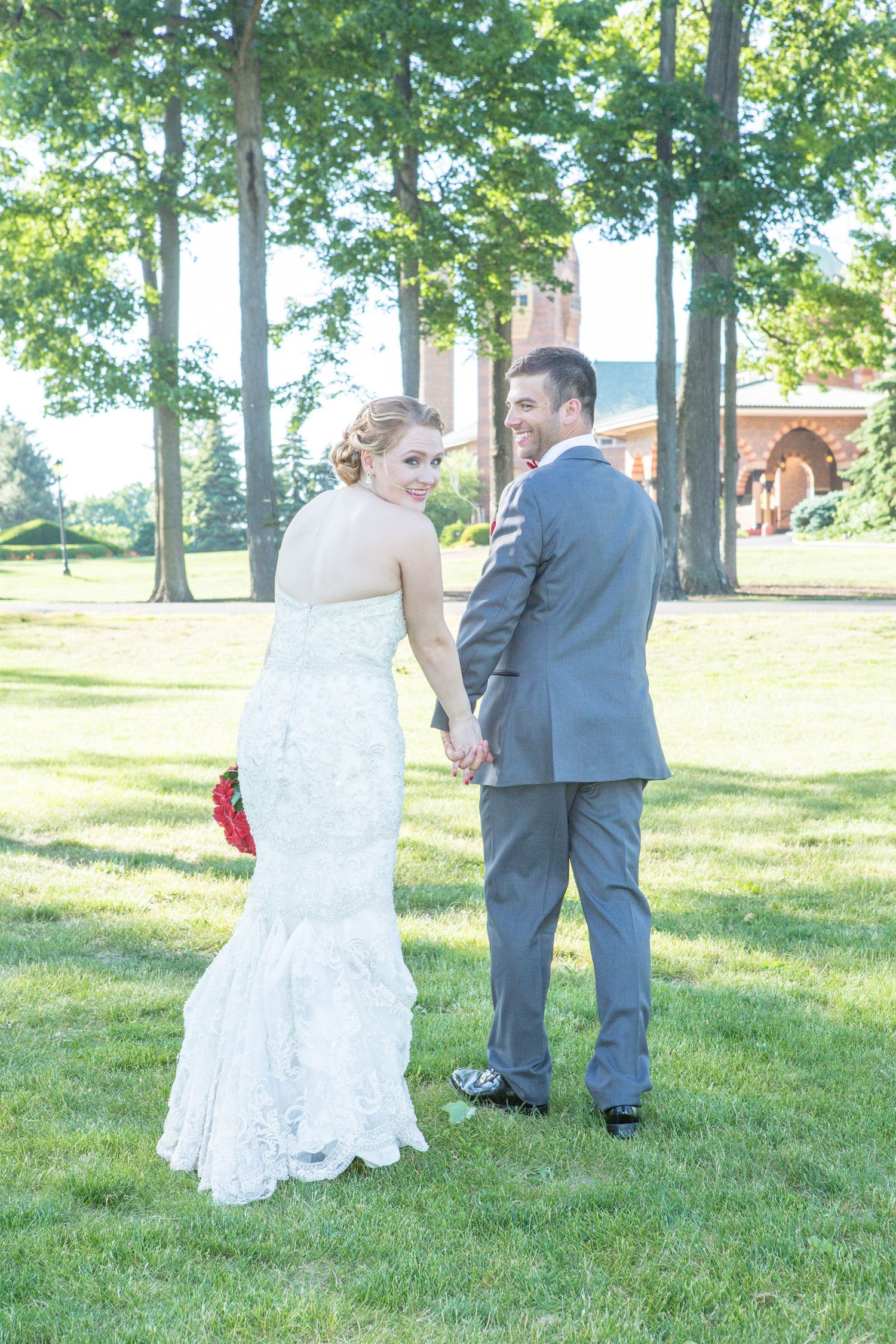 A couple stands together on a sunny lawn with trees in the background during their wedding day.