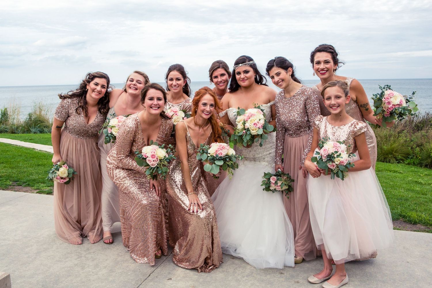 Bridal party in rose gold and blush dresses pose together on coastal overlook.