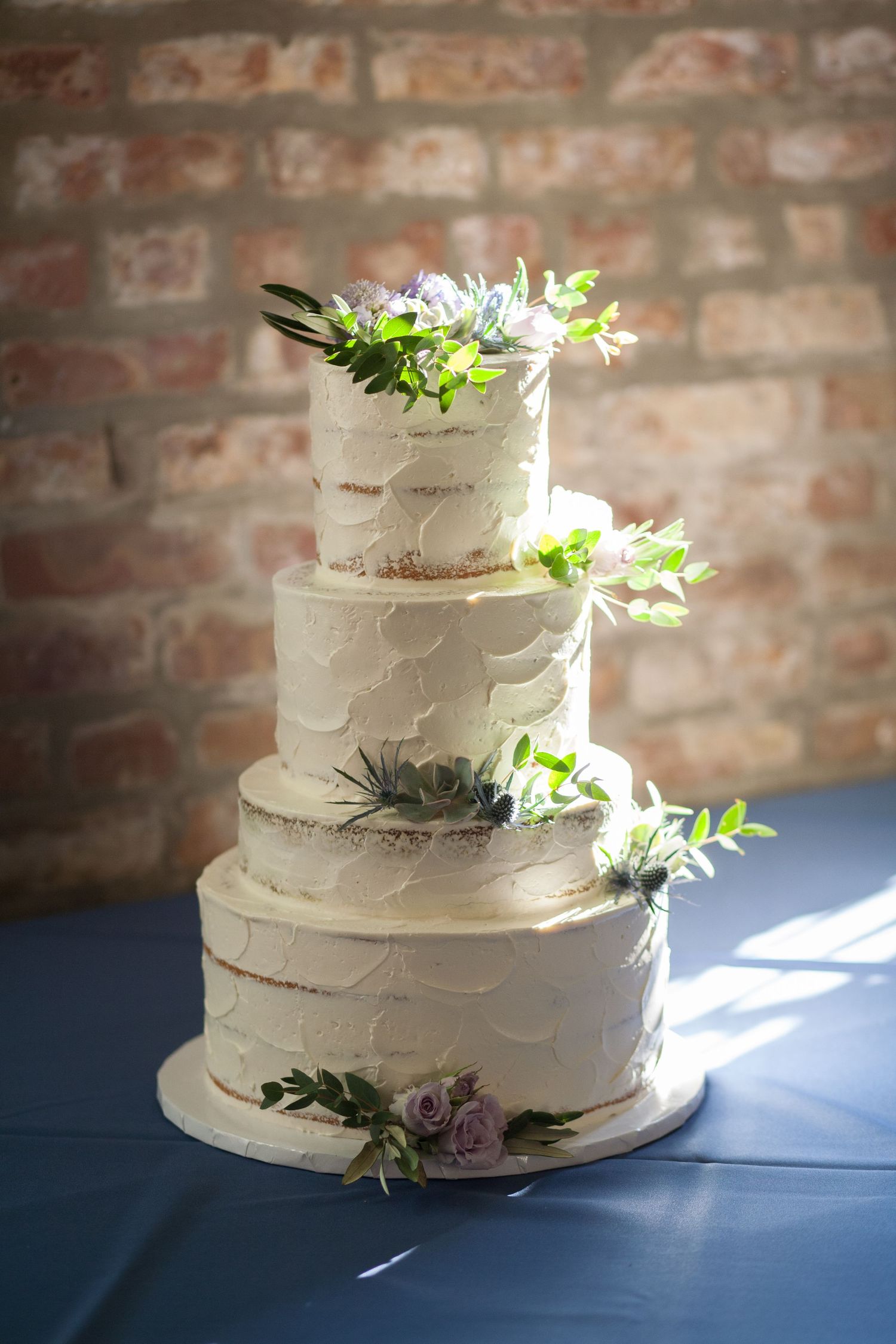 An elegant four-tier white wedding cake decorated with fresh flowers and greenery against a brick wall.