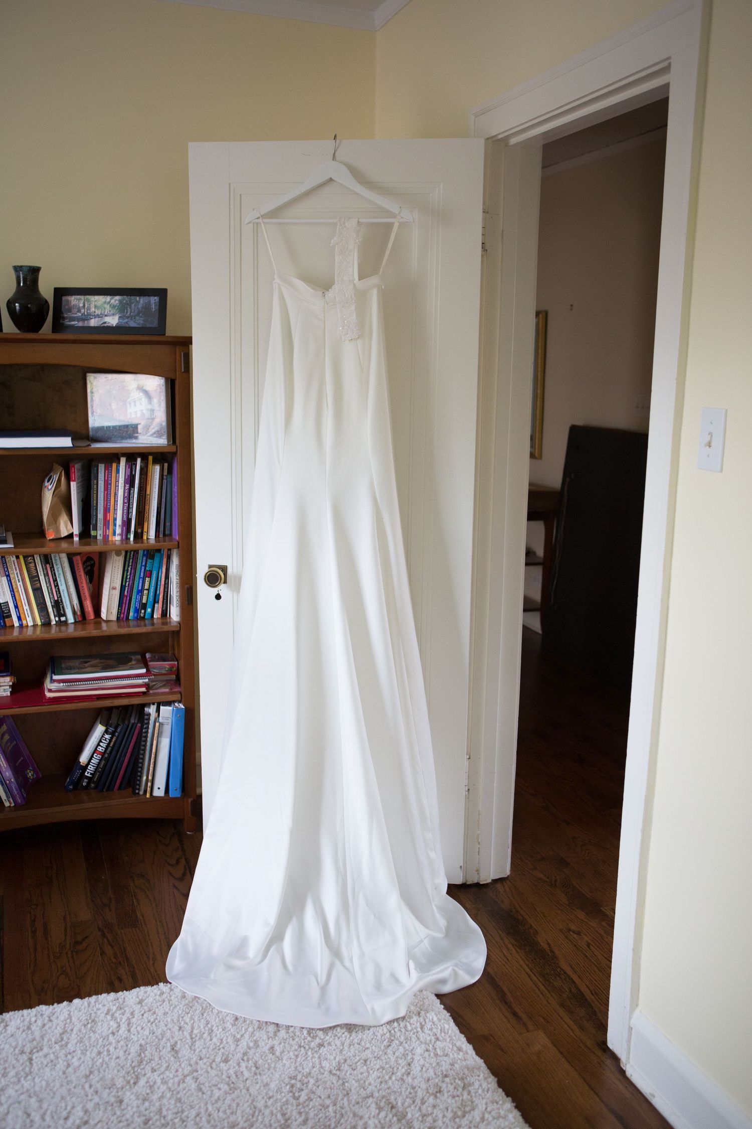 A flowing white wedding gown hangs on display against a white door near a wooden bookshelf.