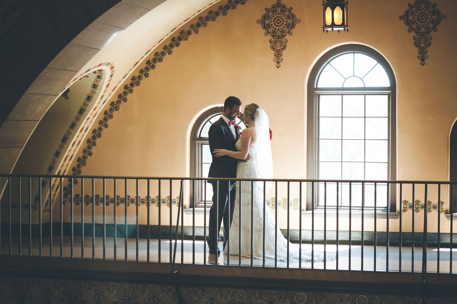 A couple shares an intimate moment on a balcony with arched windows and ornate architectural details.