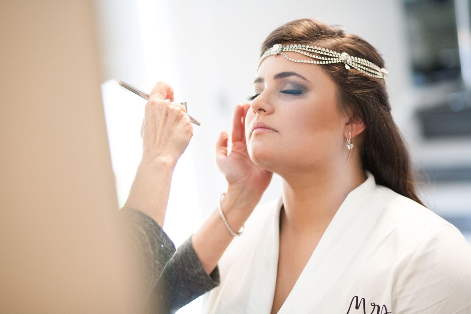 Bride getting her makeup professionally applied while wearing a white robe and crystal headpiece for her wedding day.