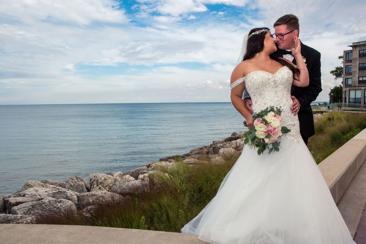 Newlyweds embrace on oceanfront terrace with bride in white strapless gown holding bouquet.