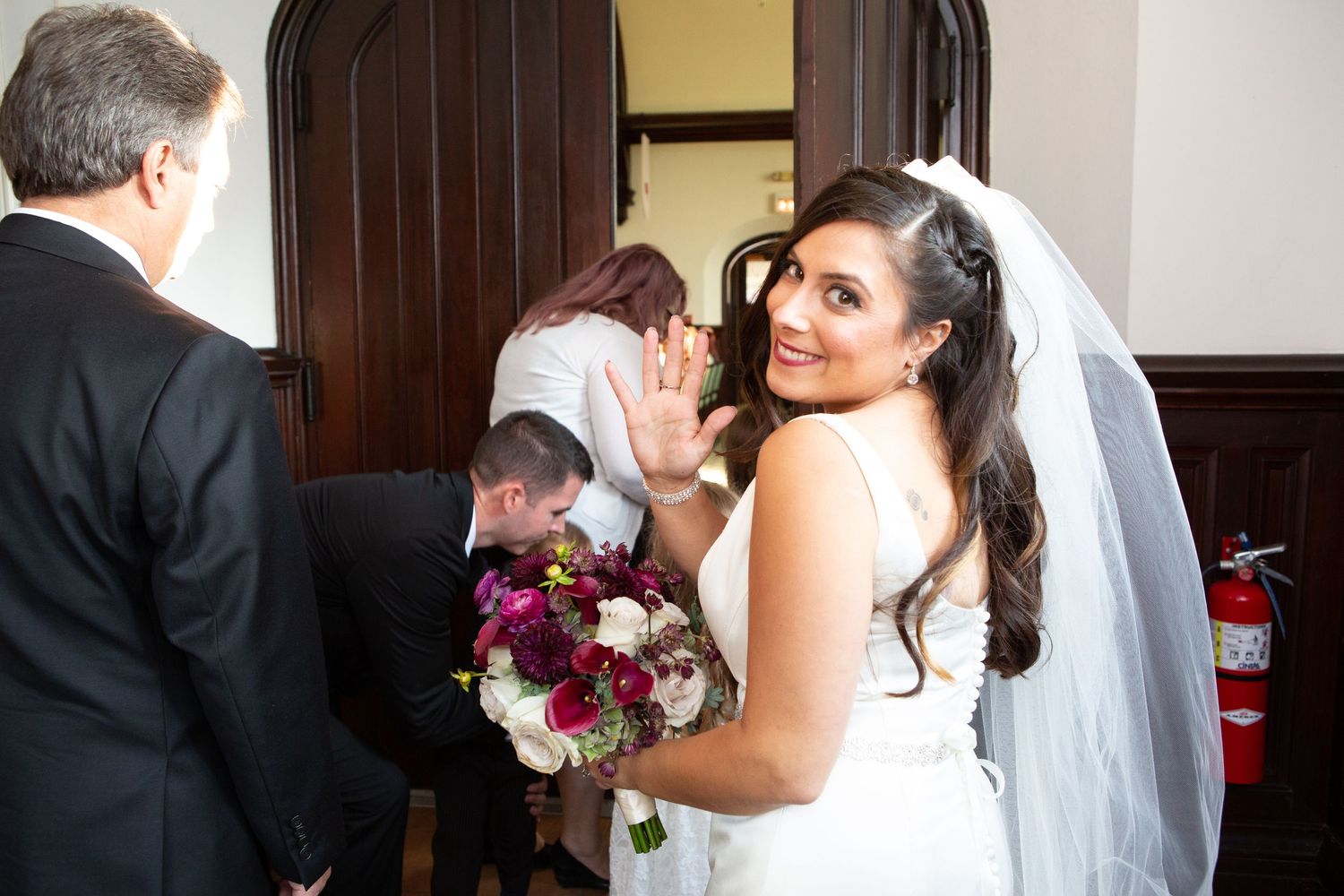 Bride with flowing veil holds purple and white bouquet while interacting with wedding guests.