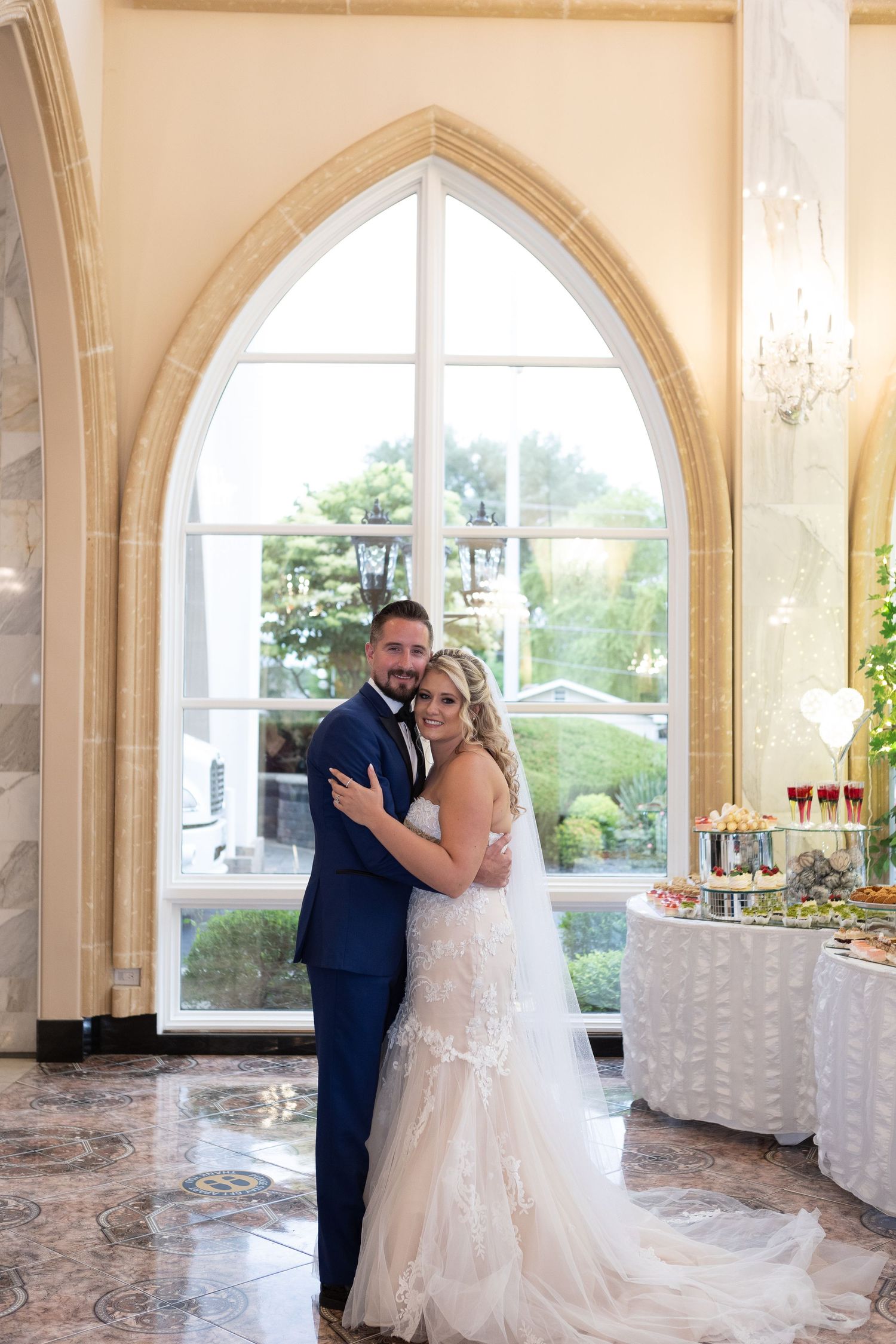 Wedding couple embracing by gothic arched window with reception table in background.