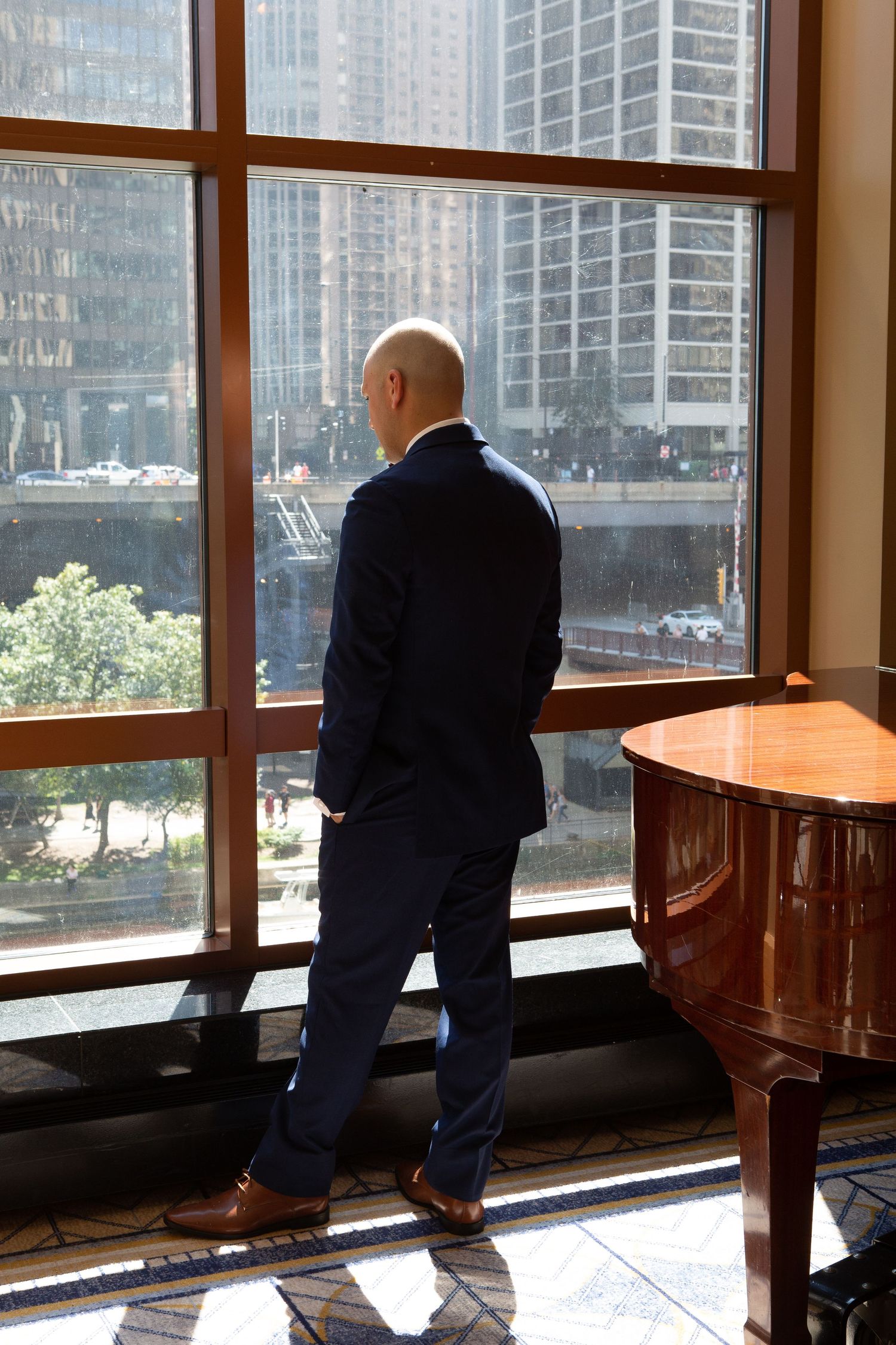 A person in a dark suit gazes out of a large window overlooking city buildings and a piano.