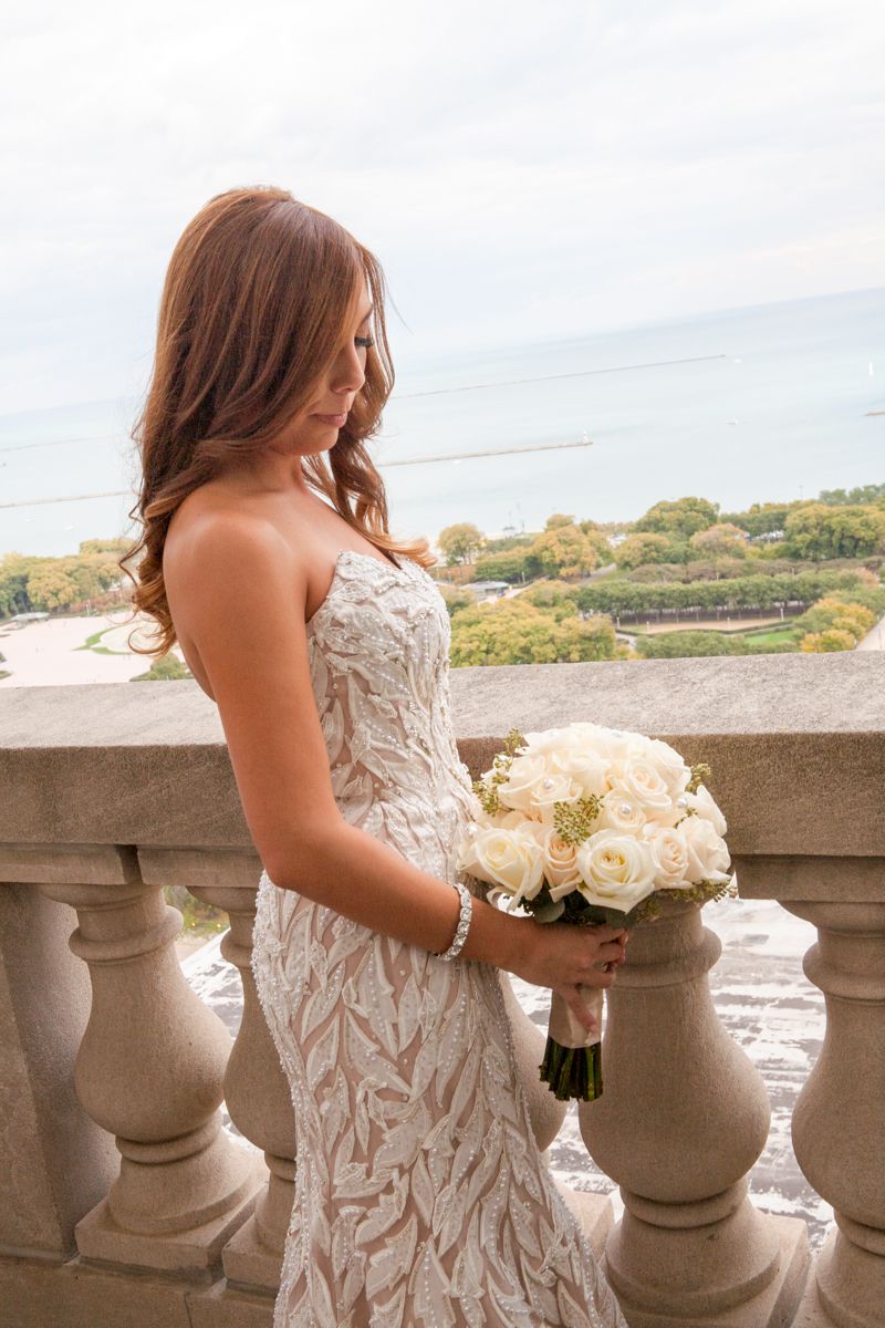 Bride in lace wedding gown holds white rose bouquet on stone balcony overlooking scenic view.