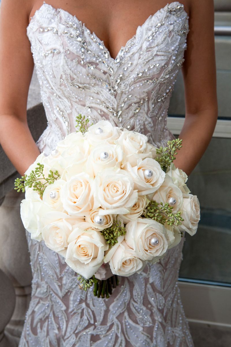 Close-up of bride holding elegant white rose bouquet with crystal-embellished wedding dress.