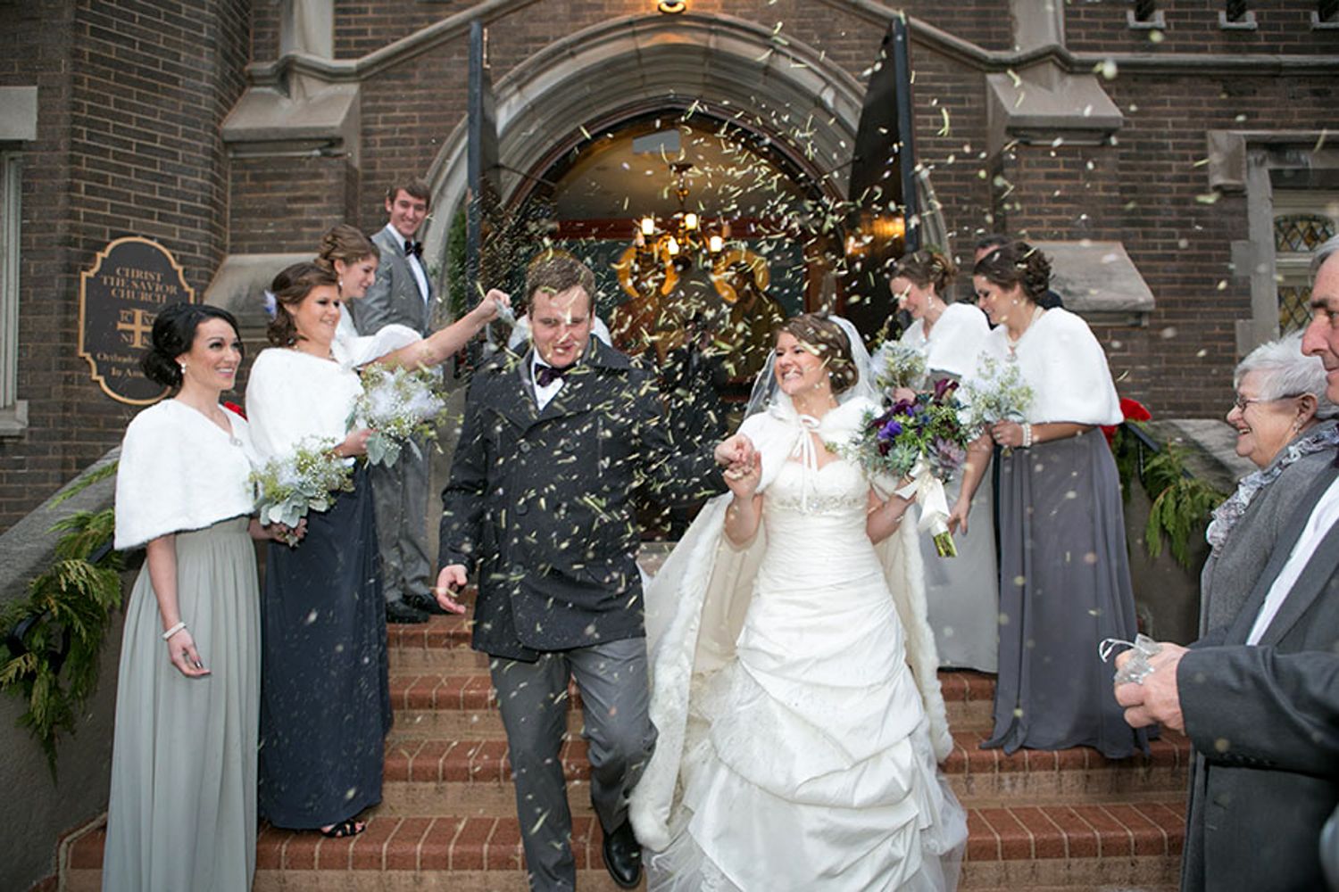 Wedding party celebrates with confetti and bubbles on brick church steps at winter ceremony.