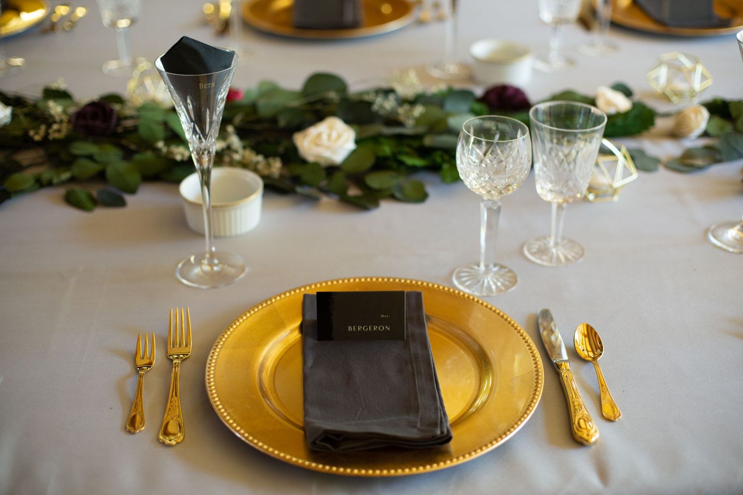 Elegant wedding place setting with gold charger plate, crystal glasses and greenery garland.