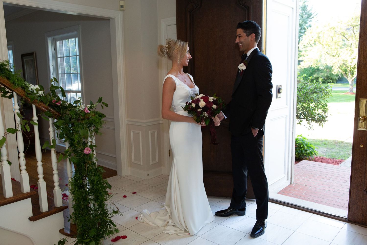 A couple in wedding attire stands near an entryway adorned with greenery garlands.