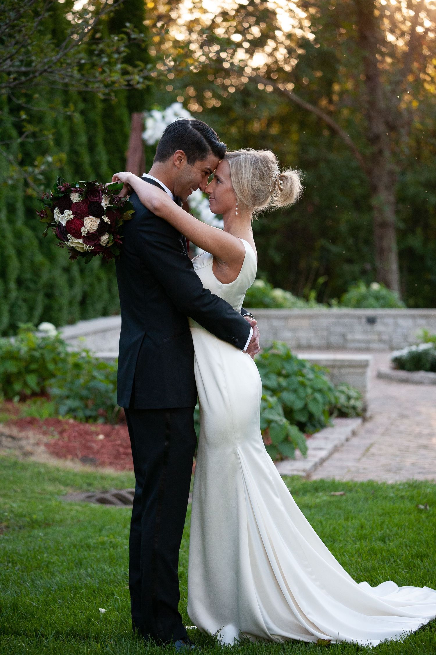 A couple shares an intimate moment outdoors in formal wedding attire with a floral bouquet.