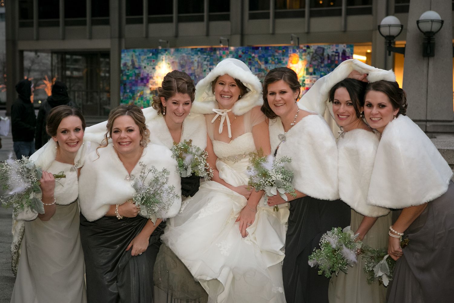 Wedding party wearing white shawls poses together on city street.