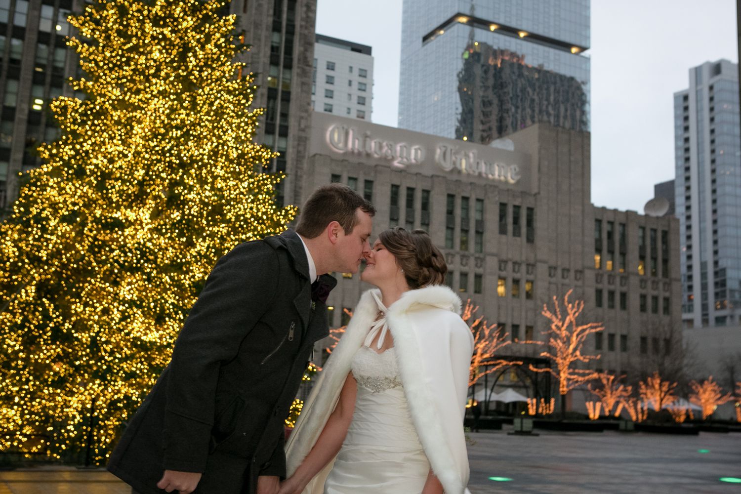 Wedding couple embraces near illuminated Christmas tree in city at night.