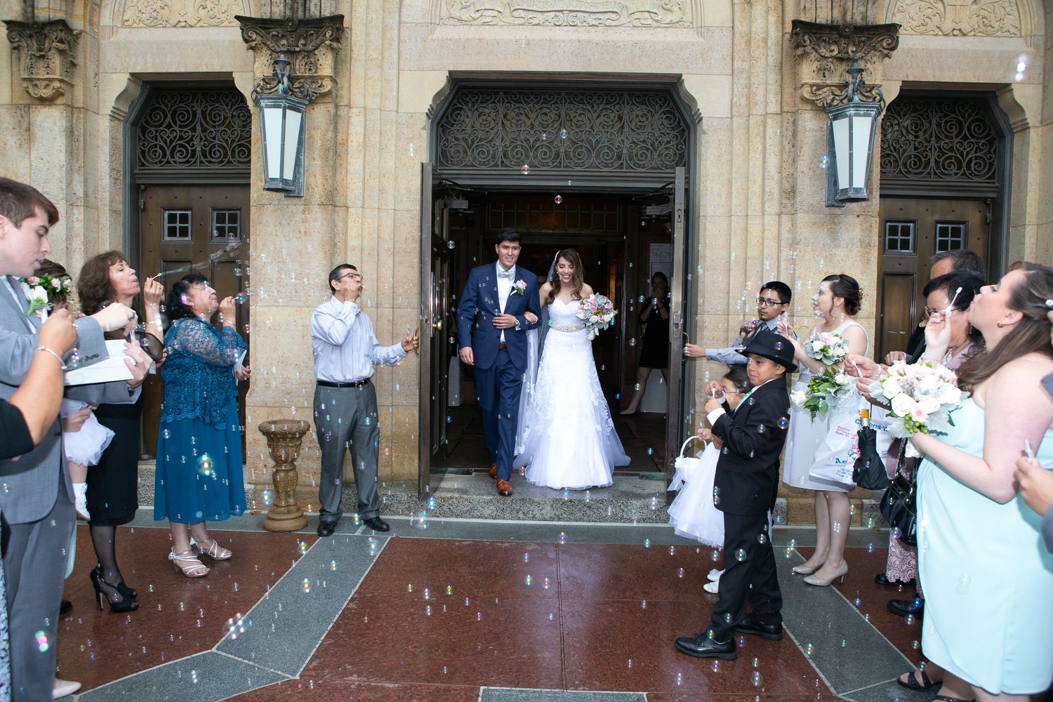 Wedding couple exits historic church entrance as guests celebrate with bubble send-off.
